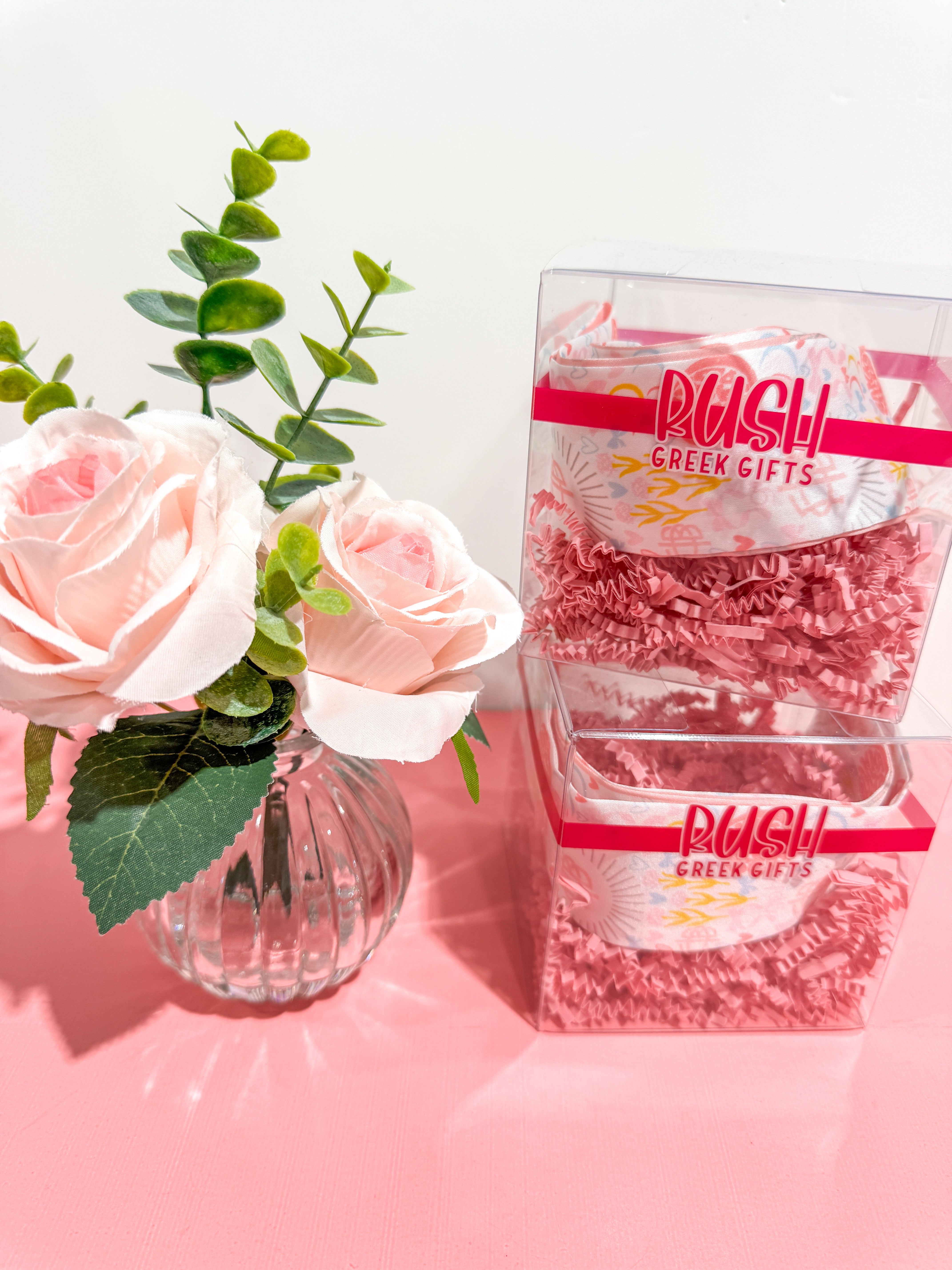 Pink roses in a vase next to a glass container with 'RUSH Greek Gifts' branding on a pink surface.