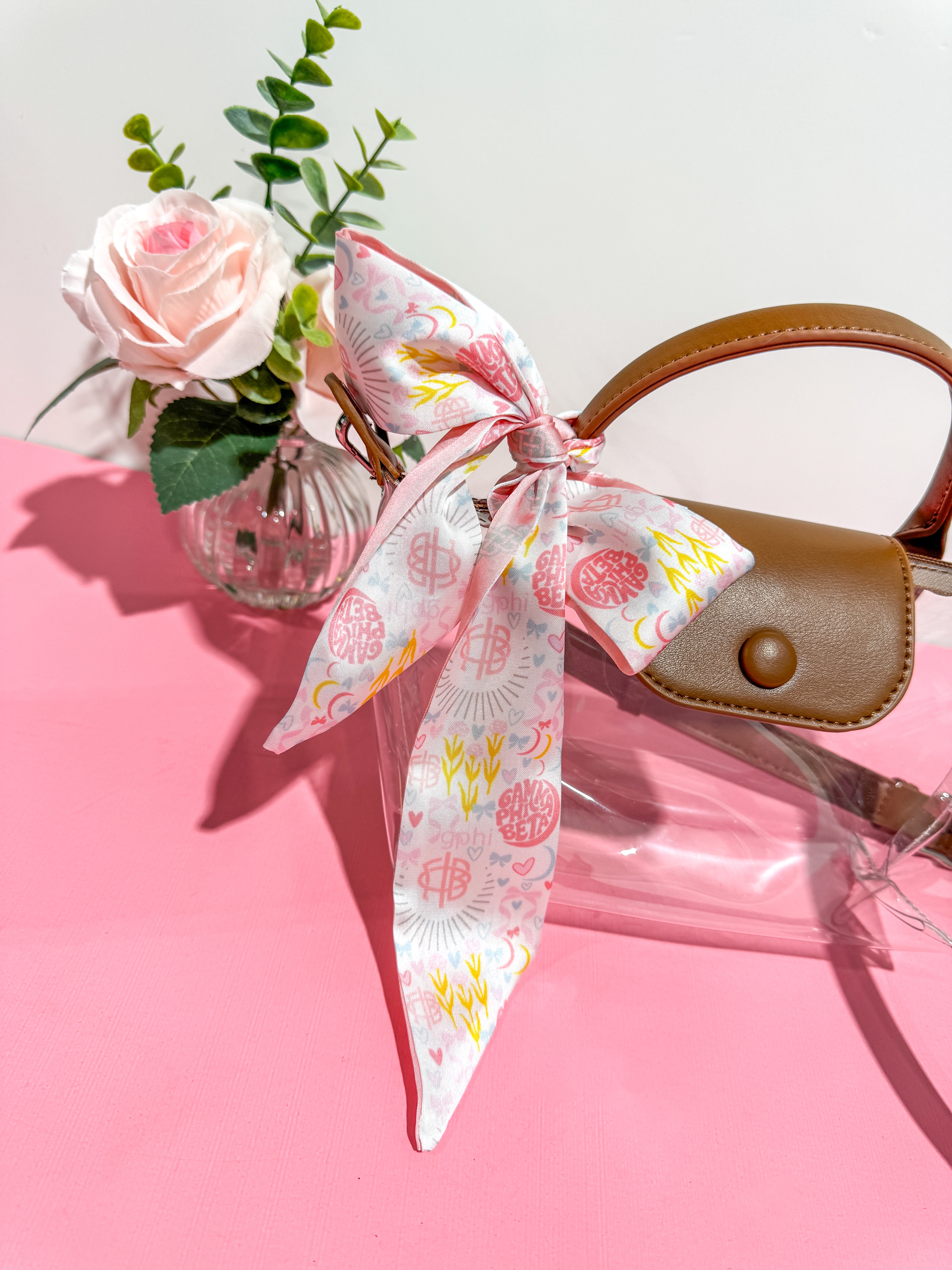 Handbag with a floral sorority silky tie on a pink surface with a vase of flowers.