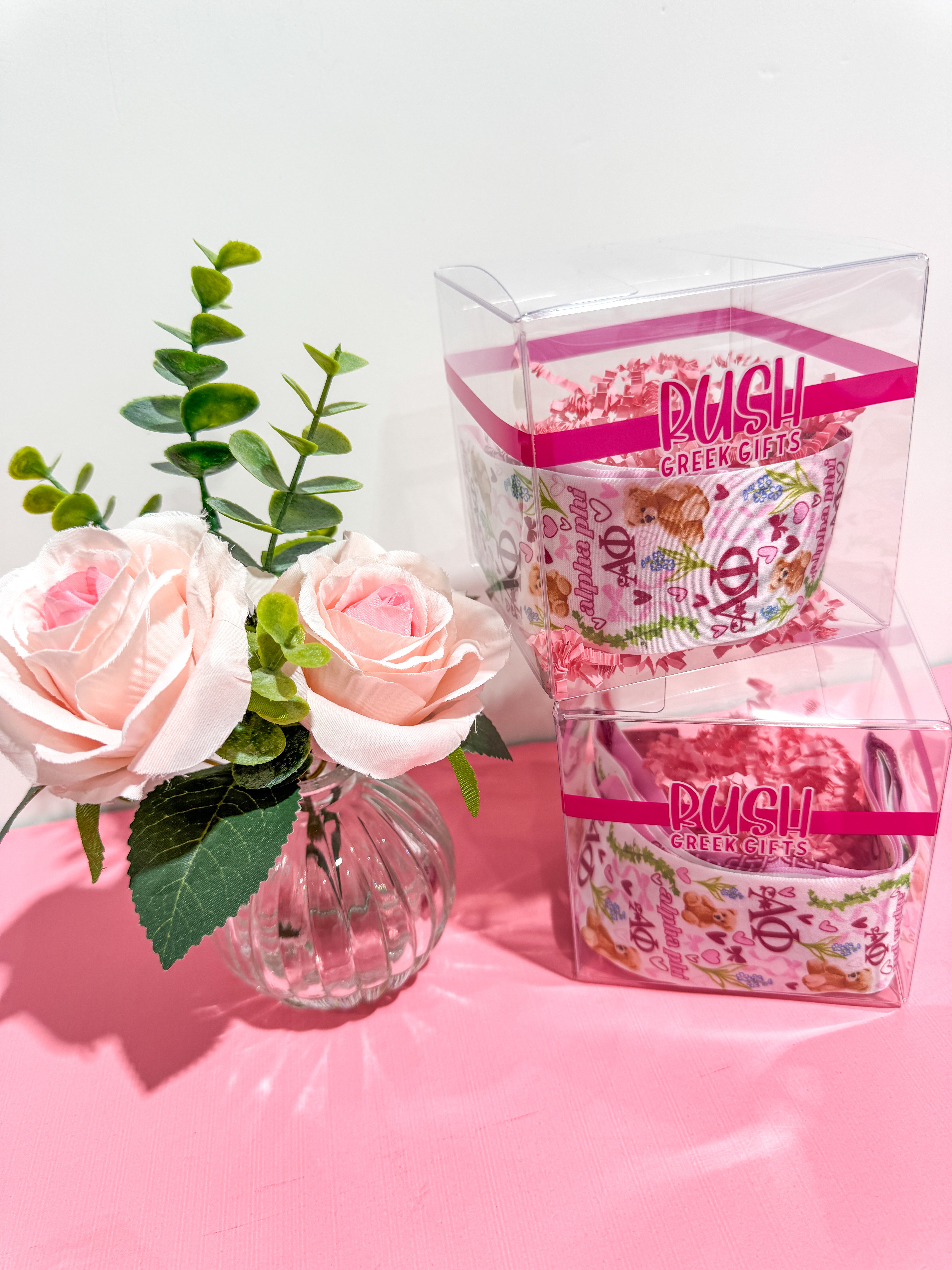 Two retail packages with floral designs on a pink surface with pink roses and green leaves for Alpha Phi sorority. 