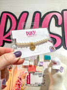 Pearl bracelet with gold Alpha Chi Omega charm in clear packaging held by a hand, with 'Rush Greek Gifts' branding in the background.